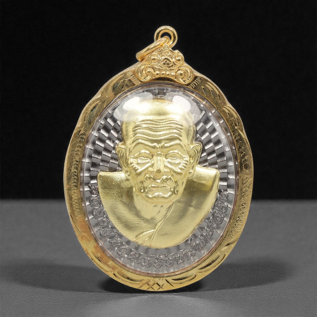 A Thai Bi-Metallic Monk Portrait Amulet with Gilt-Metal Frame, 21st Century, 65FP: The amulet is of oval form, featuring a high-relief gilt bust portrait of a Buddhist monk on the obverse, set against a silver-toned metal ground with vertical radiating linear motifs; incised Thai sc