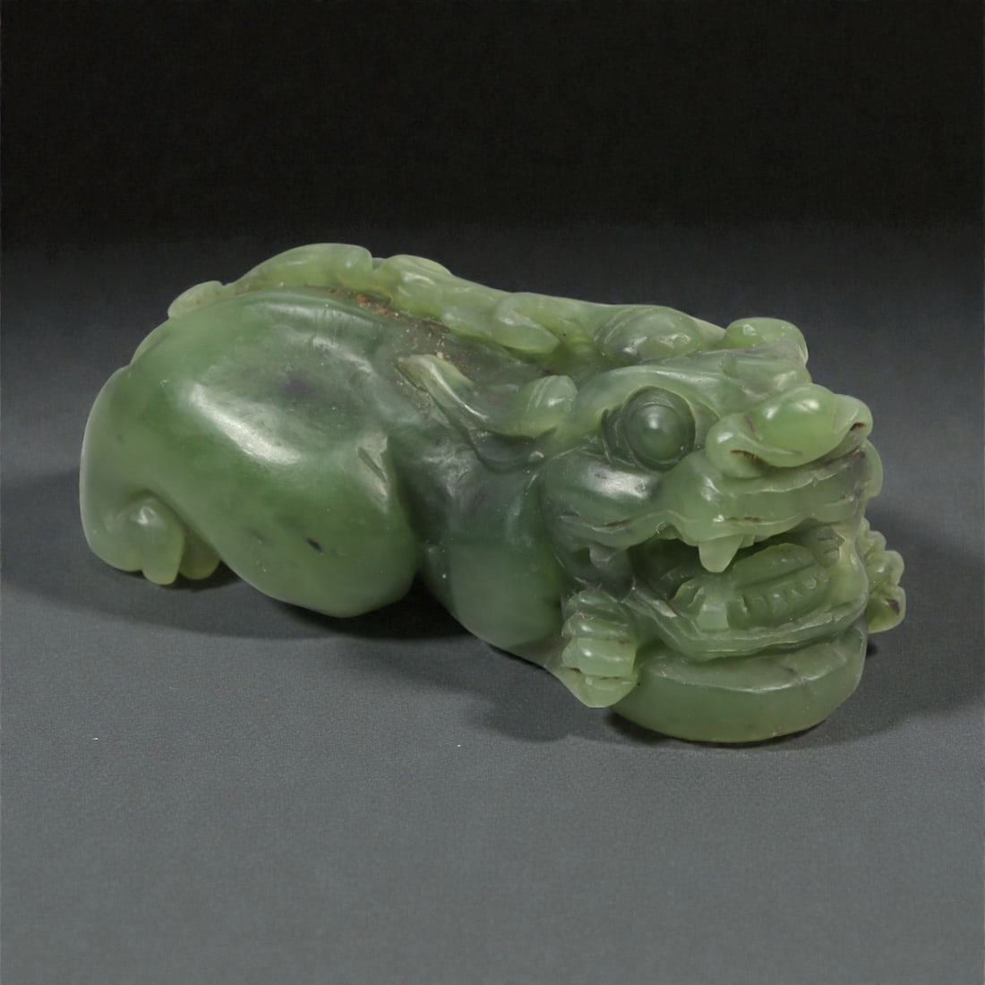 A Chinese Nephrite Jade Mythical Beast Figure, 20th Century, 4YA: Carved from dark green nephrite jade with areas of lighter translucency and scattered natural inclusions, the figure represents a recumbent mythical beast shown in a compact, crouching posture. The he