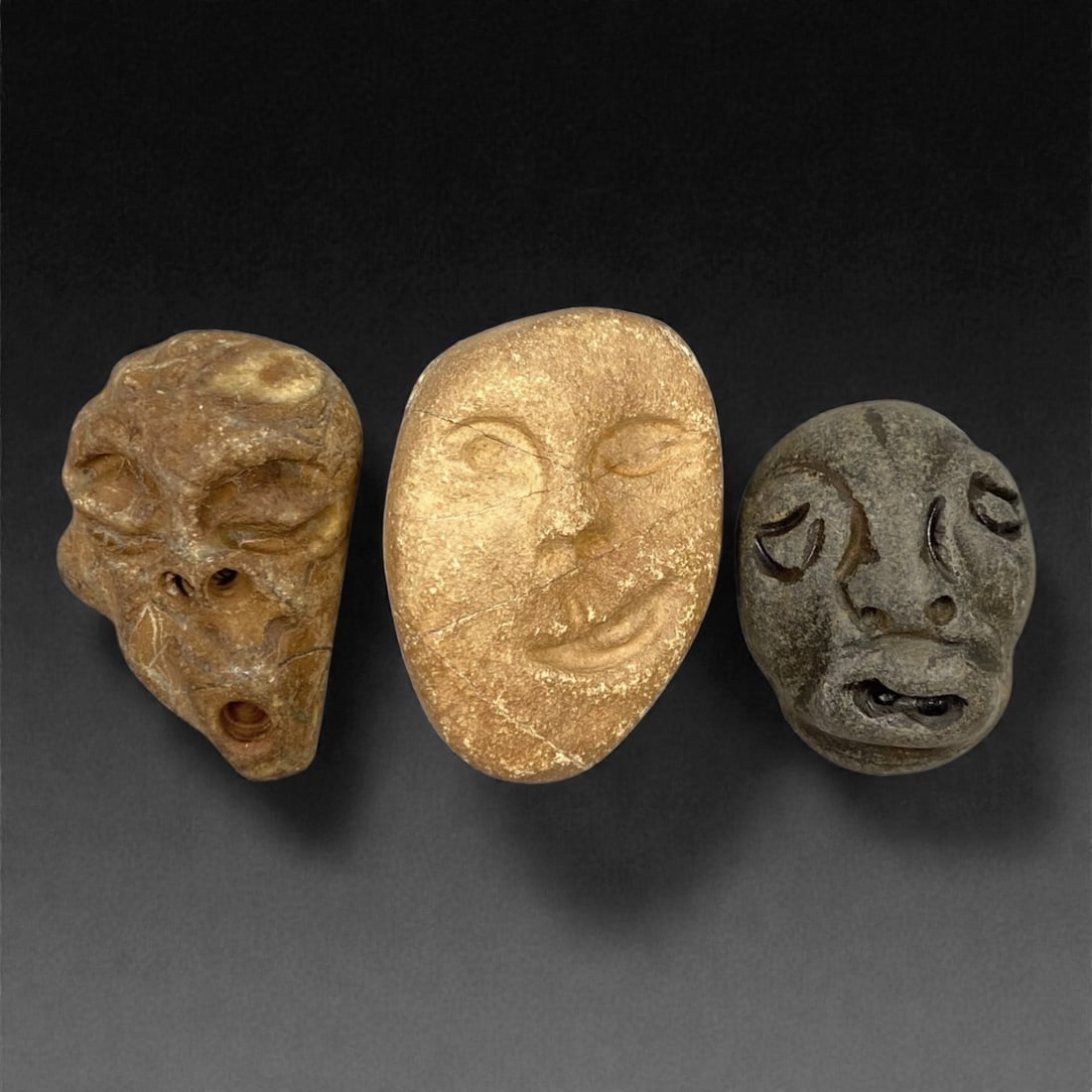 A Group of Three Medieval Stone Maskette Amulets, 10th-14th Century, 50FP: The group comprises three stone maskette amulets, each carved from distinct stone types with varied surface treatments. The first is a brown stone mask with an irregular, angular form, featuring reces