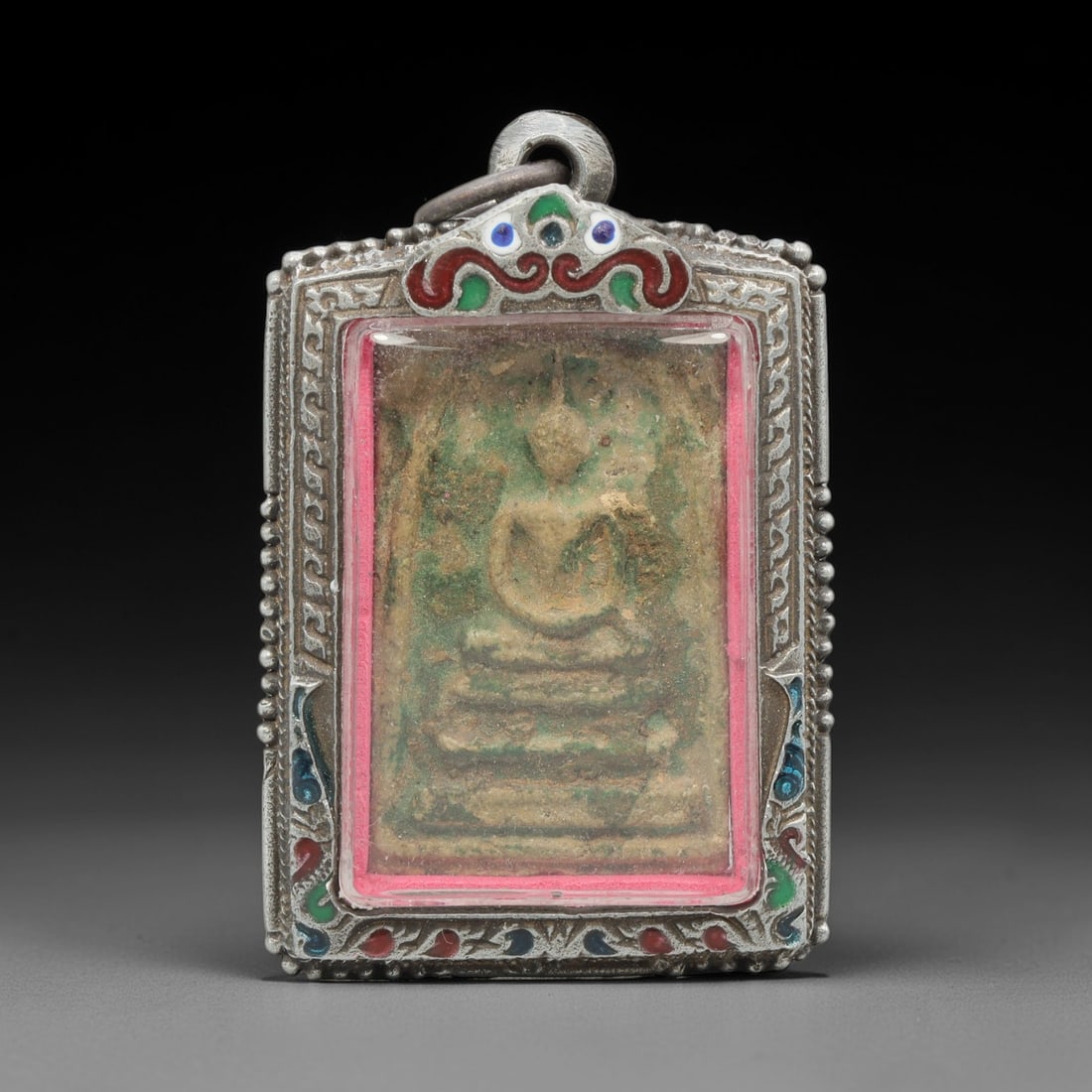 A Thai Buddha Amulet in Enameled Metal Case, Late 20th Century, 82ZH: A rectangular Buddha amulet, measuring 5.5 x 3.5 cm, housed in a hinged metal case with red, green, and blue enamel inlays. The obverse of the case displays a seated Buddha figure in relief, set withi