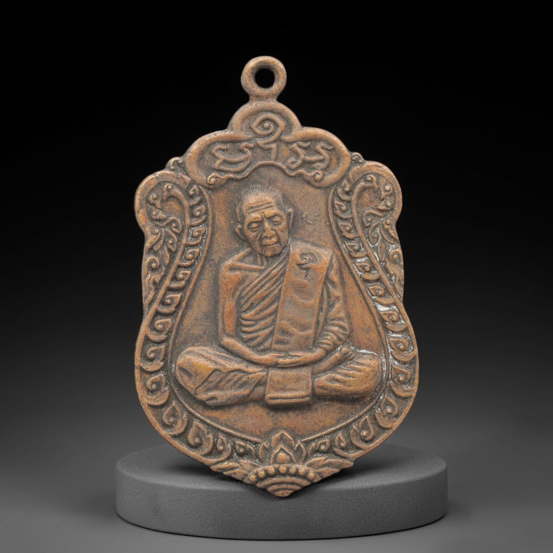 A Thai Copper Alloy Buddhist Amulet, Late 20th Century, 98FP: A shield-shaped copper alloy amulet, measuring 4.2 x 2.7 cm. The obverse depicts a seated Buddhist monk in meditative mudra, framed by ornate scrollwork and a lotus motif at the base. The reverse feat