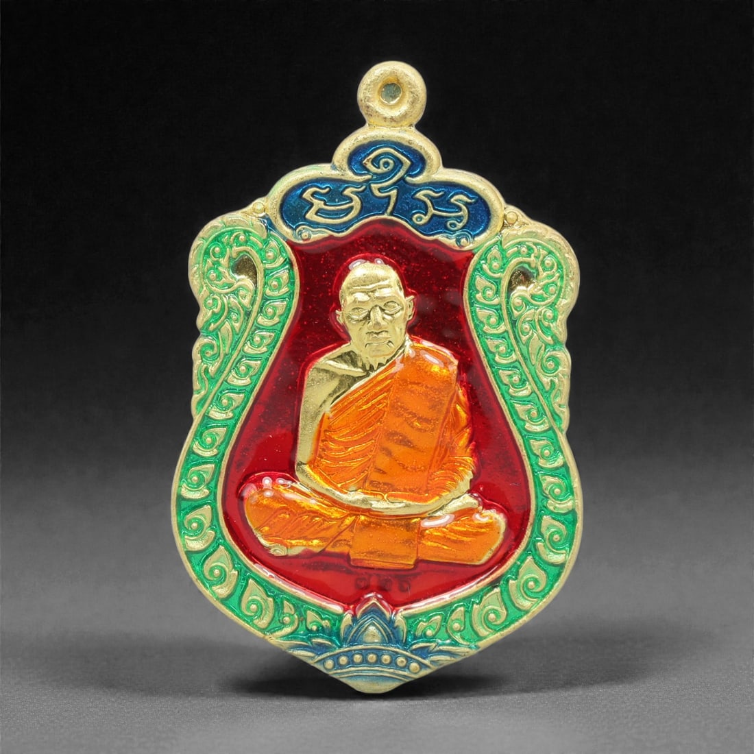 A Thai Gilt-Metal and Enamel Monk Portrait Amulet, 21st Century, 70FP: Cast from metal with a gilt surface finish, the amulet has a shield-shaped form with an integral suspension loop at the apex. The obverse features a high-relief gilt bust portrait of a seated Buddhist