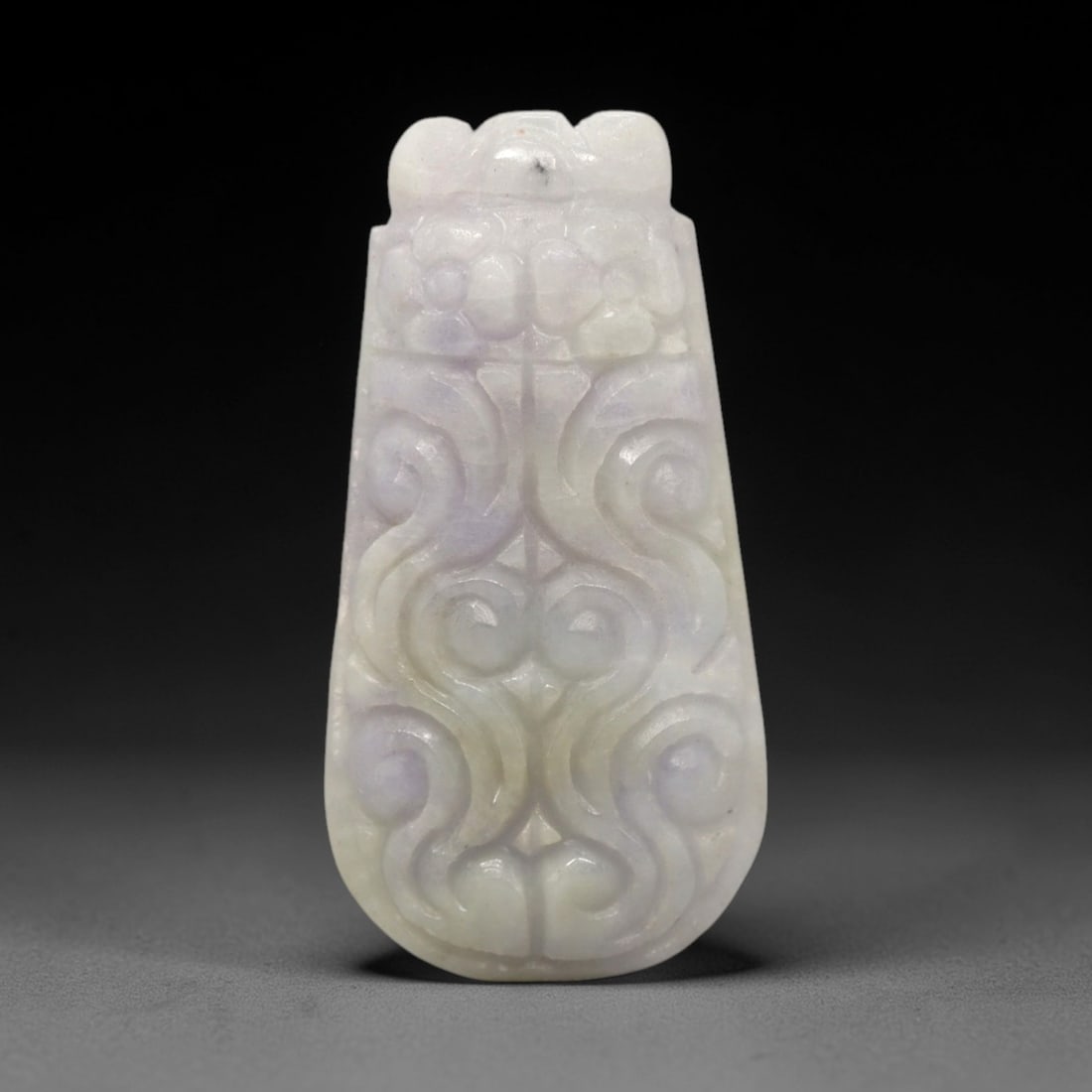 A Chinese Jadeite Jade Scrollwork Pendant, 20th Century, 83FR: Carved from translucent jadeite jade with prominent pale lavender tonal variations and minor white inclusions, the pendant has a tapered, shield-like form with a stylized double-loop bail at the apex.