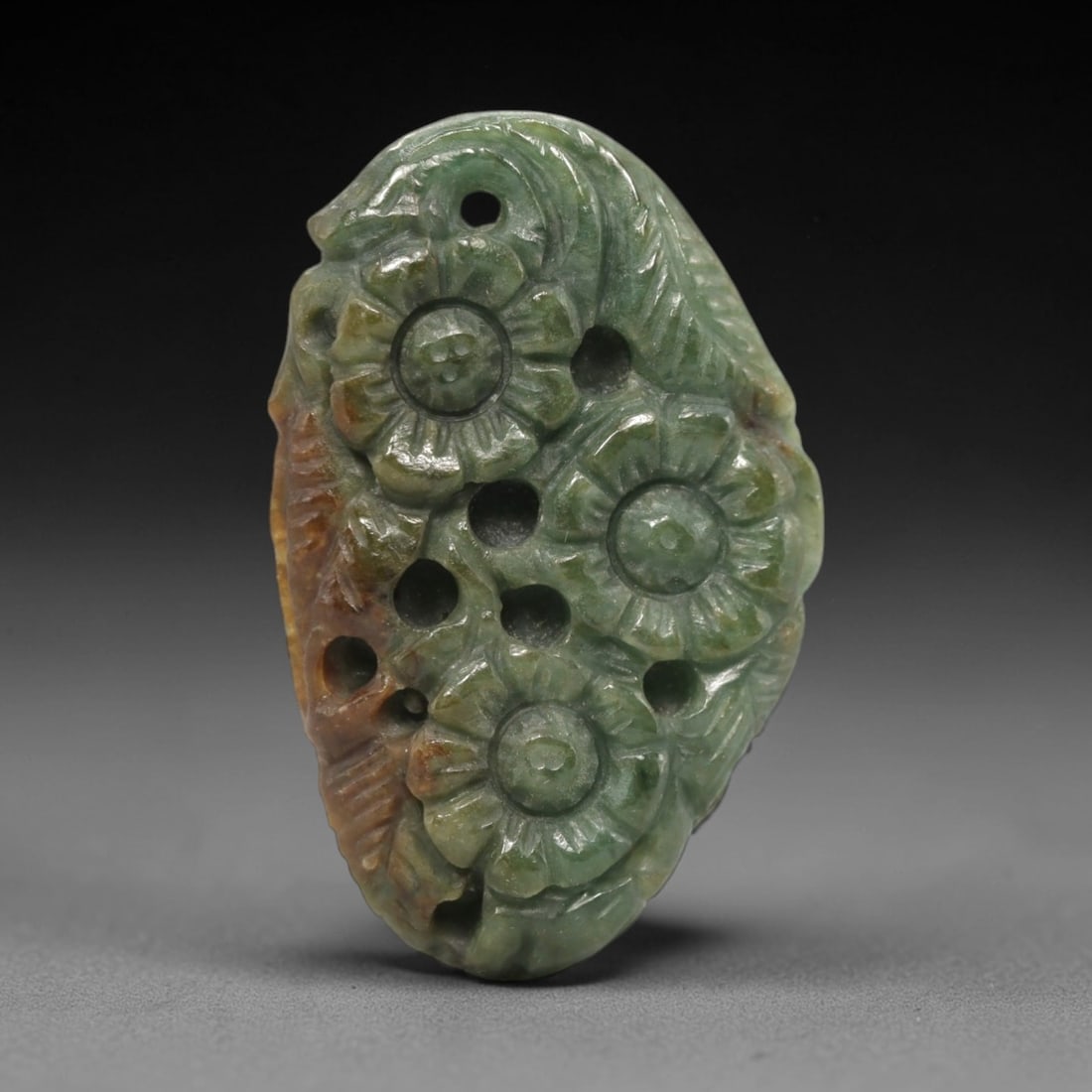 A Chinese Jadeite Jade Floral Pendant, 20th Century, 87FR: Carved from variegated green and brownish jadeite jade with natural tonal variations, the pendant is irregularly shaped with a drilled suspension hole at the top. It is decorated with openwork carving