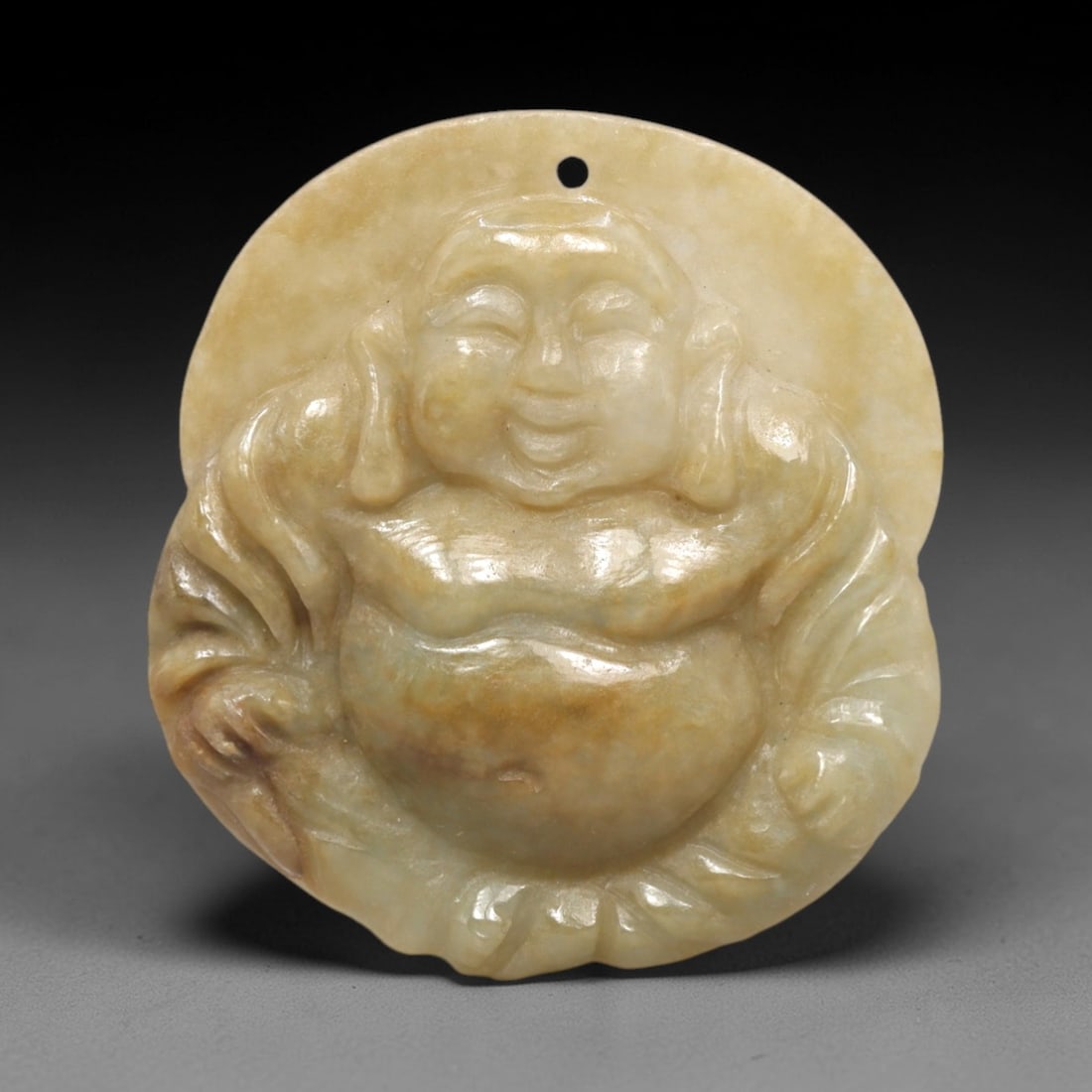 A Chinese Jadeite Jade Budai Pendant, 20th Century, 98FR: Carved from pale yellowish jadeite jade with natural tonal variations and subtle inclusions, the pendant is irregularly circular in form, with a drilled suspension hole at the top. It depicts Budai se