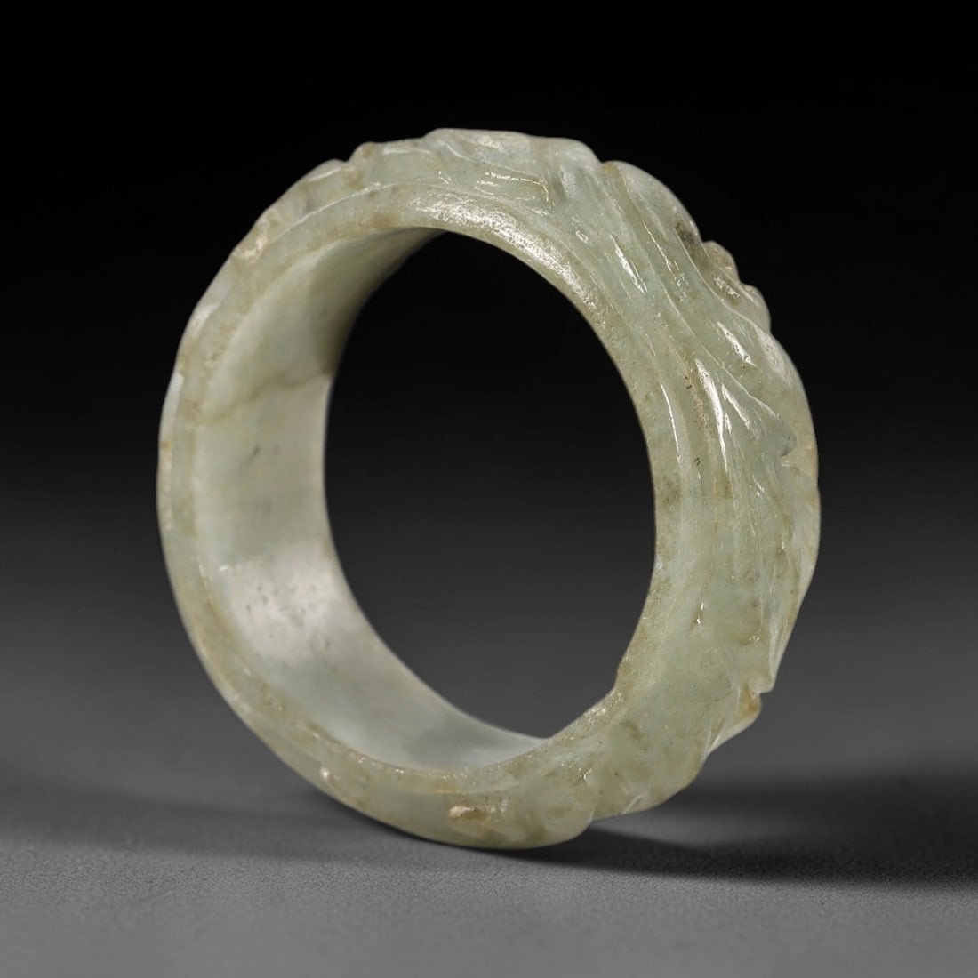 A Chinese Jadeite Jade Floral-Form Ring, Qing Dynasty, 62FS: Carved from pale greenish-white jadeite jade with natural diffuse veining and subtle internal inclusions, the object is formed as a circular ring with a continuous relief carving of floral and foliate
