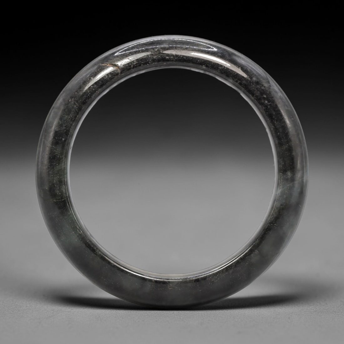 A Burmese Jadeite Jade Bangle, 20th Century, 11FS: A Burmese jadeite jade bangle, with a dark grey matrix and subtle lighter grey mineral inclusions. The bangle is of circular form, with a smooth, polished surface revealing the internal crystalline st