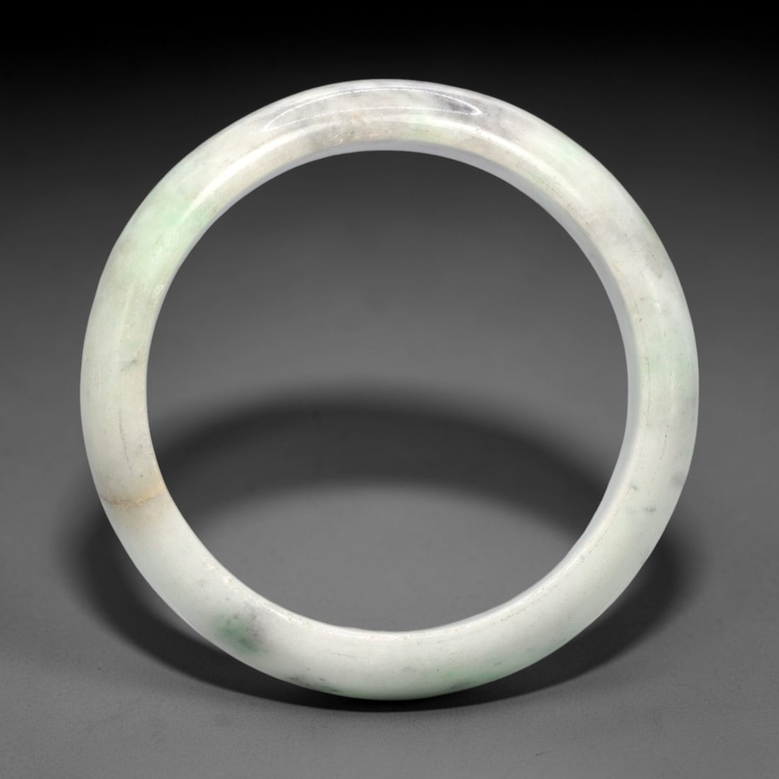 A Burmese Jadeite Jade Bangle, 20th Century, 17FS: A Burmese jadeite jade bangle, with a pale white matrix and scattered light green mineral inclusions. The bangle is of circular form, with a smooth, polished surface revealing the internal crystalline