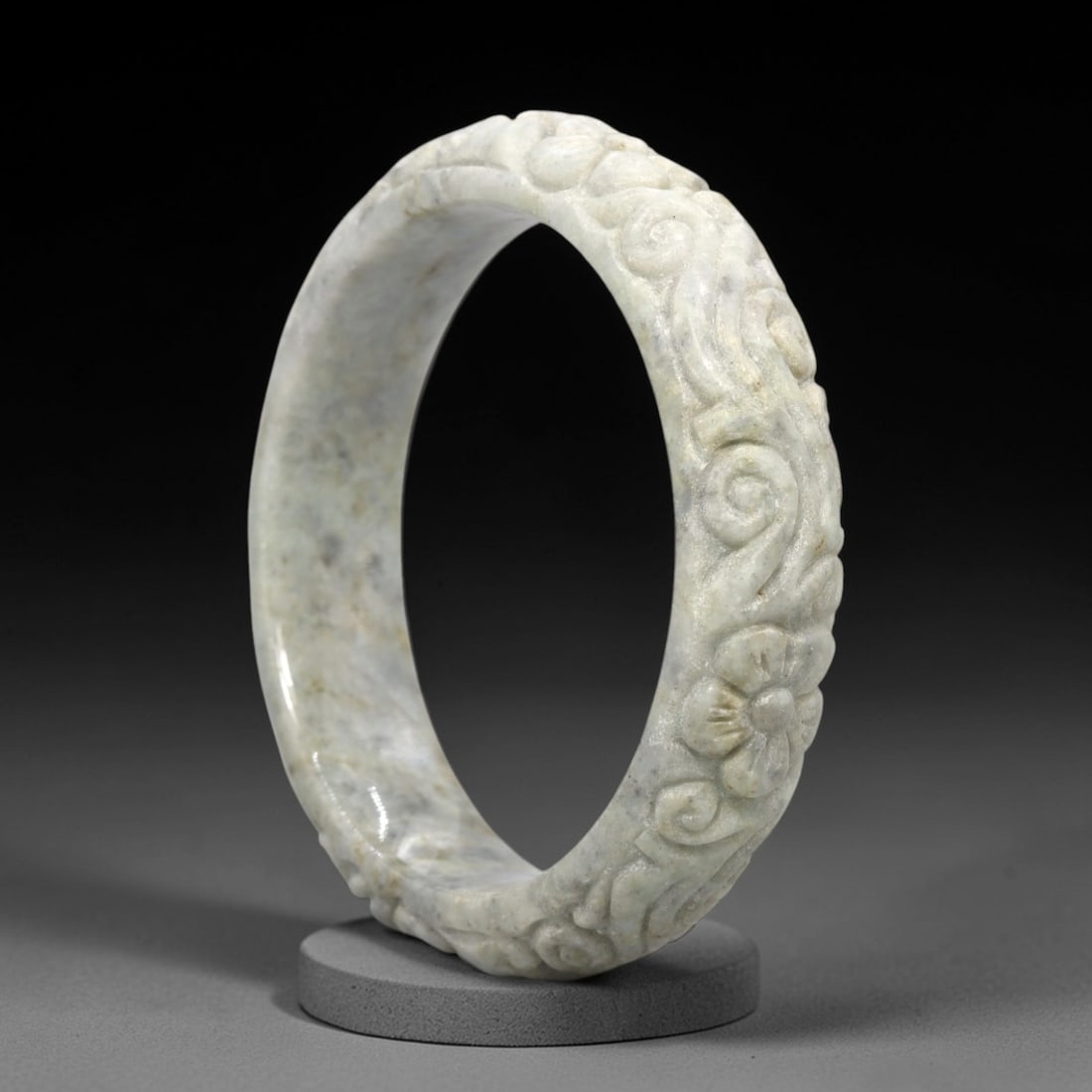 A Chinese Jadeite Jade Bangle Bracelet, 20th Century, 30FS: Carved from pale greyish-white jadeite jade with natural tonal variation and subtle surface texture, the bangle is formed as a continuous circular band. The exterior surface is decorated with raised r