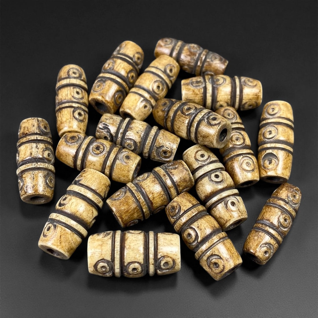 A Group of Tibetan Bone Beads, 20th Century, 1FT: The group comprises seventeen cylindrical beads, carved from bone with a light tan base and dark incised decoration. Each bead is segmented by horizontal bands, with circular motifs incised into the r