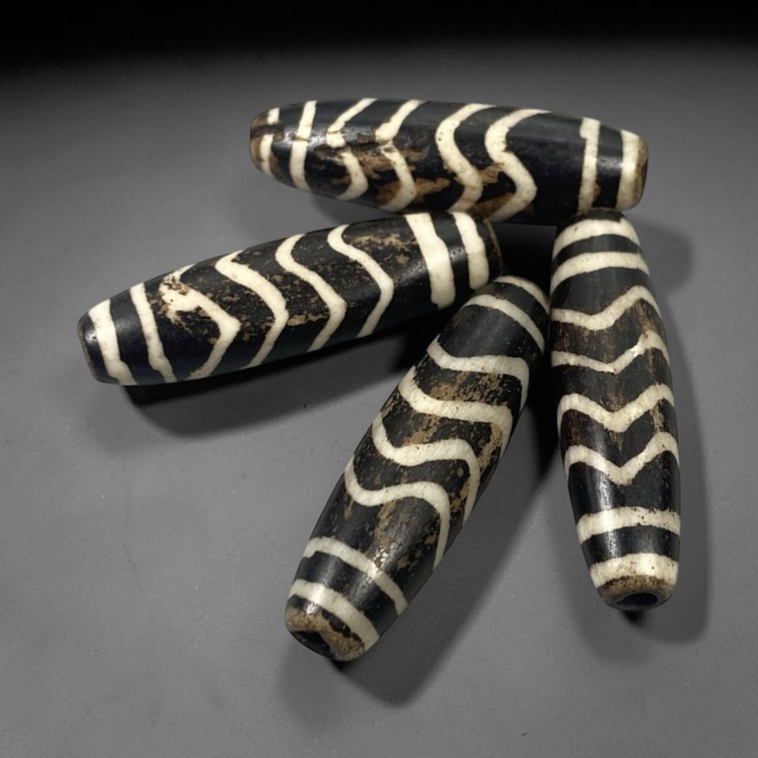 A Group of Burmese Pumtek Wood Beads, 19th-20th Century, 15FT: The group consists of four elongated cylindrical beads, carved from wood and treated with a dark surface, overlaid with white geometric patterns. The beads display repeating zigzag and linear motifs,