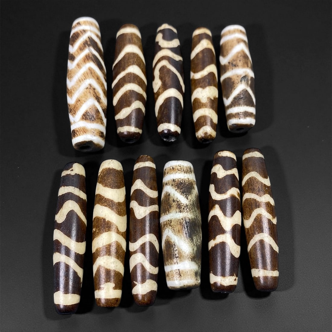 A Group of Burmese Pumtek Wood Beads, 19th-20th Century, 2FT: The group consists of eleven cylindrical beads, carved from wood and treated with a dark surface, overlaid with white and light tan geometric patterns. The beads display a range of consistent motifs,