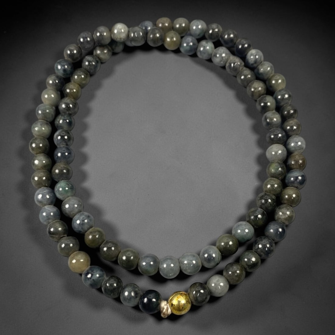 A Chinese Jadeite Jade Bead Necklace, 20th Century, 62ZK: Composed of spherical beads of dark grey and green jadeite jade with natural veining and subtle tonal variations, the necklace is strung in a continuous loop. The beads exhibit a smooth, polished surf