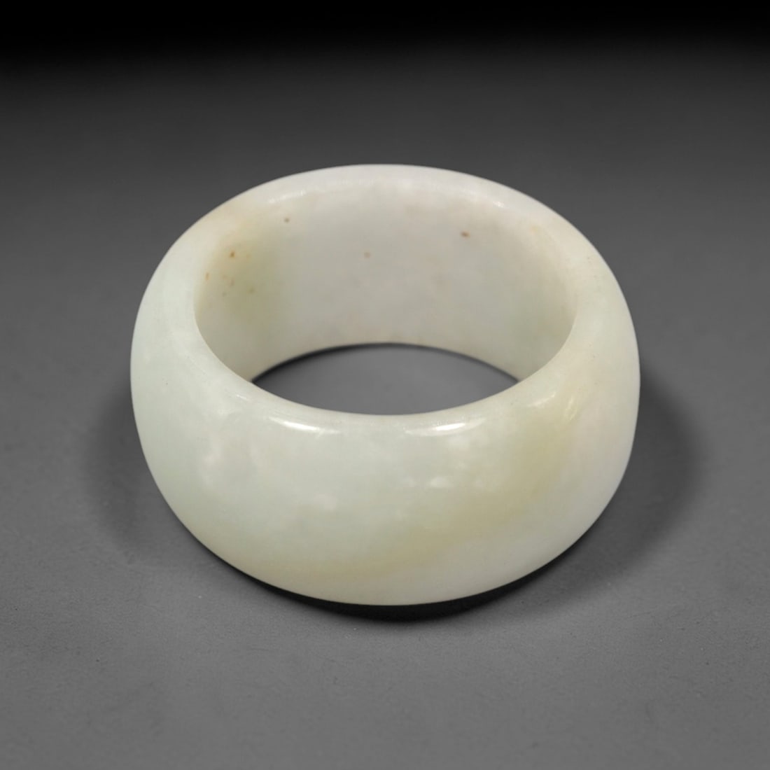 A Chinese Jadeite Jade Ring, 20th Century, 25FT: Carved from pale greenish-white jadeite jade with subtle tonal variation and minor natural inclusions, the ring is formed as a thick, circular band with a smooth, highly polished surface. The form is