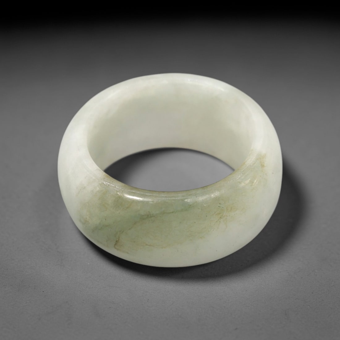 A Chinese Jadeite Jade Ring, 20th Century, 26FT: Carved from pale white jadeite jade with natural greenish inclusions and subtle tonal variation, the ring is formed as a thick, circular band with a smooth, highly polished surface. The form is robust