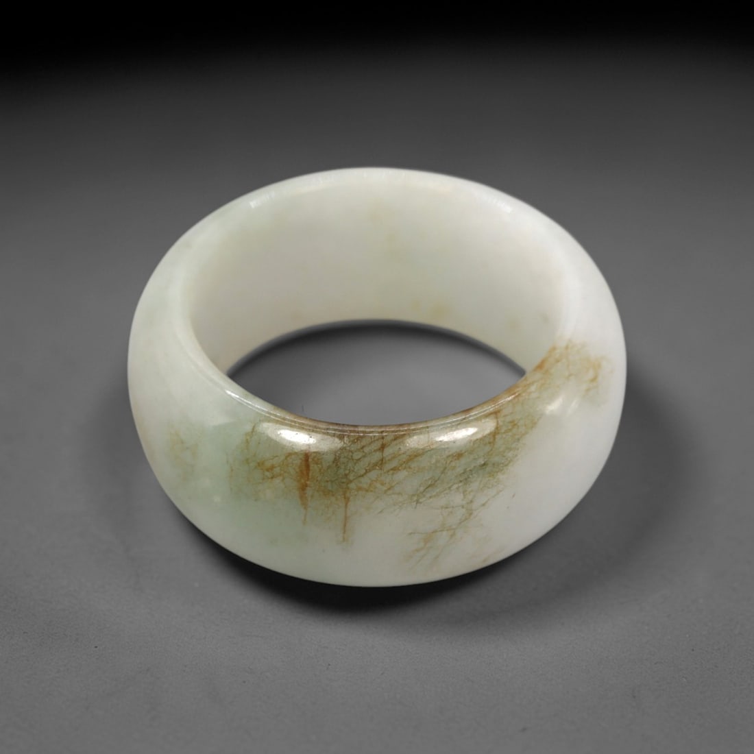 A Chinese Jadeite Jade Ring, 20th Century, 29FT: Carved from pale white jadeite jade with natural green and brownish inclusions and subtle tonal variation, the ring is formed as a thick, circular band with a smooth, highly polished surface. The form