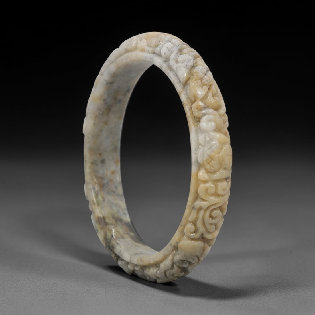 A Chinese Carved Jadeite Jade Bangle Bracelet, 20th Century, 50ZL: A fine jadeite bangle of flattened circular form, masterfully carved in relief with a continuous band of scrolling foliate and auspicious cloud motifs. The stone exhibits a striking variegated palette