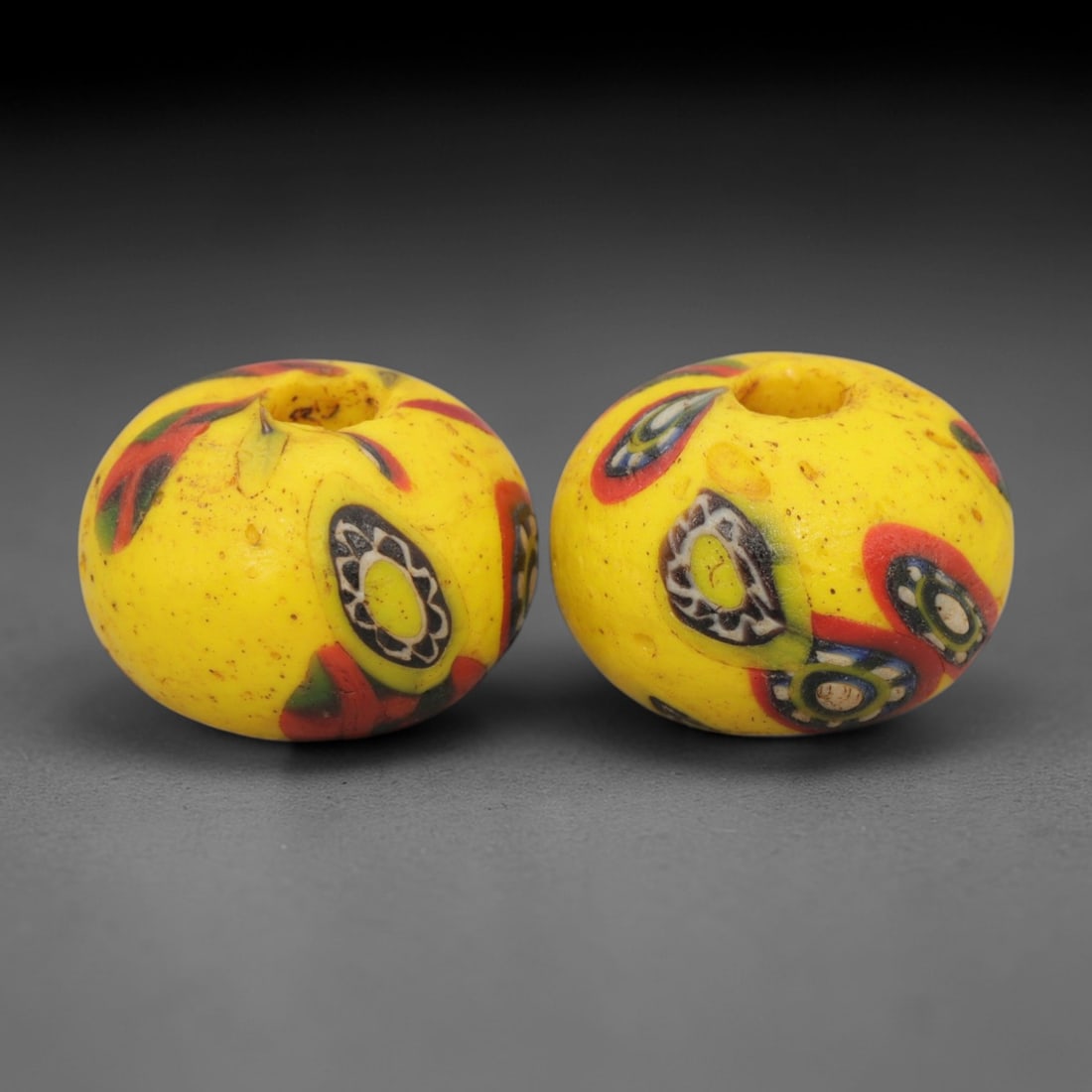 A Pair of Venetian Millefiori Glass Beads, 19th-20th Century, 64AD: Fabricated from opaque yellow glass, each bead is formed as a sphere with a central drilled perforation. The surface is decorated with characteristic millefiori canes, featuring circular eye-like moti