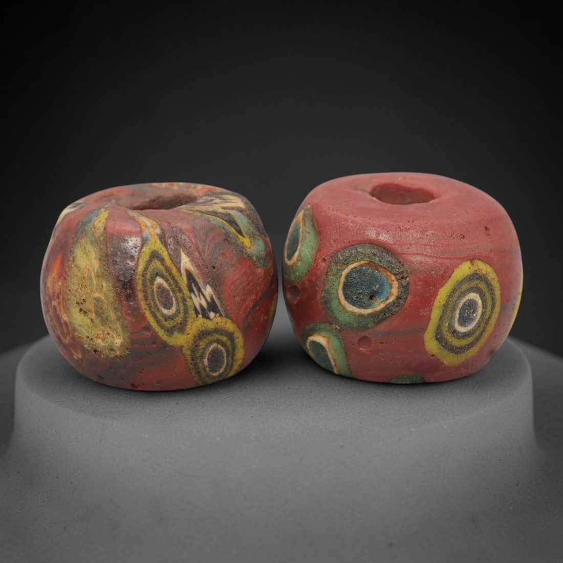 A Pair of Venetian Millefiori Glass Beads, 19th-20th Century, 53AD: Fabricated from opaque red glass, each bead is formed as a short cylinder with a central drilled perforation. The surface is decorated with characteristic millefiori canes: one bead displays circular