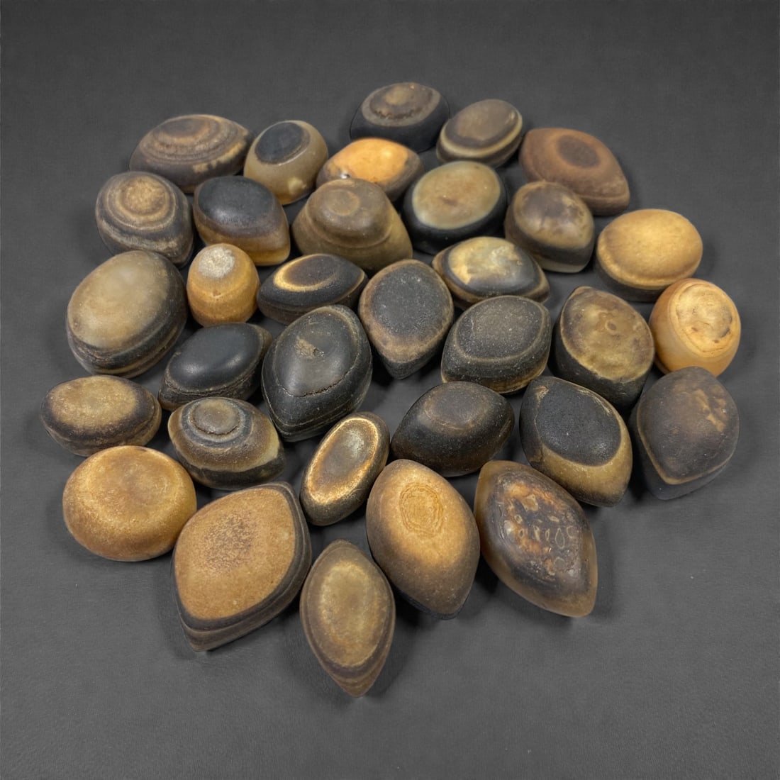 A Group of Tibetan Luk Mik ("Goat's Eye") Agate Beads, 11th-14th Century CE, 86V: Comprising thirty-three agate beads of ovoid, lenticular, and slightly irregular form, carved from naturally banded agate in tones of amber, honey, brown, grey, and black, the group is distinguished b