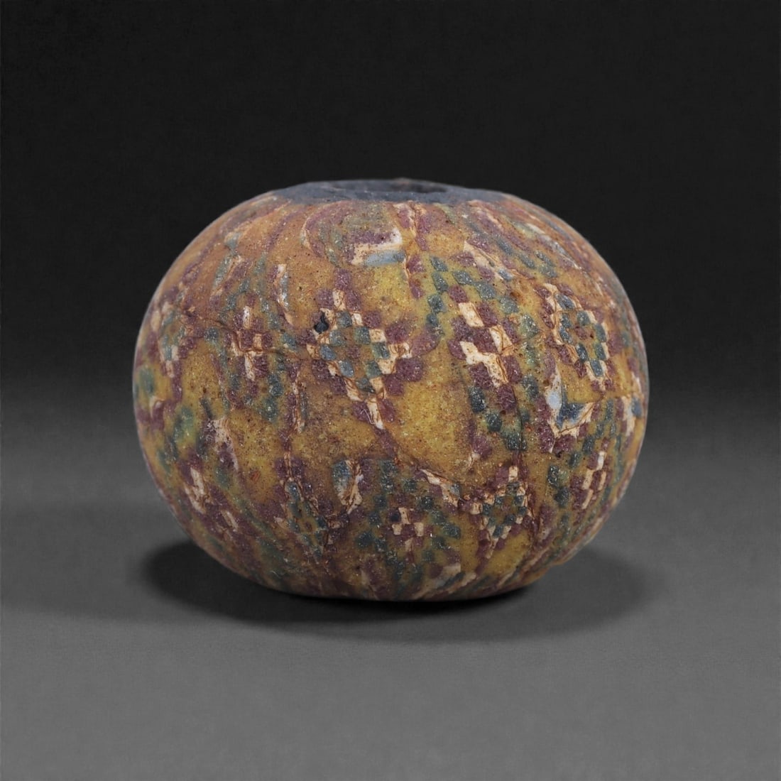 Ancient Glass Bead - Near Eastern Mosaic Ornament, 62J: Composed of multicolored mosaic glass, this small bead is formed by carefully layered canes fused into a compact spherical body. The exterior presents a dense arrangement of checker and lozenge motifs