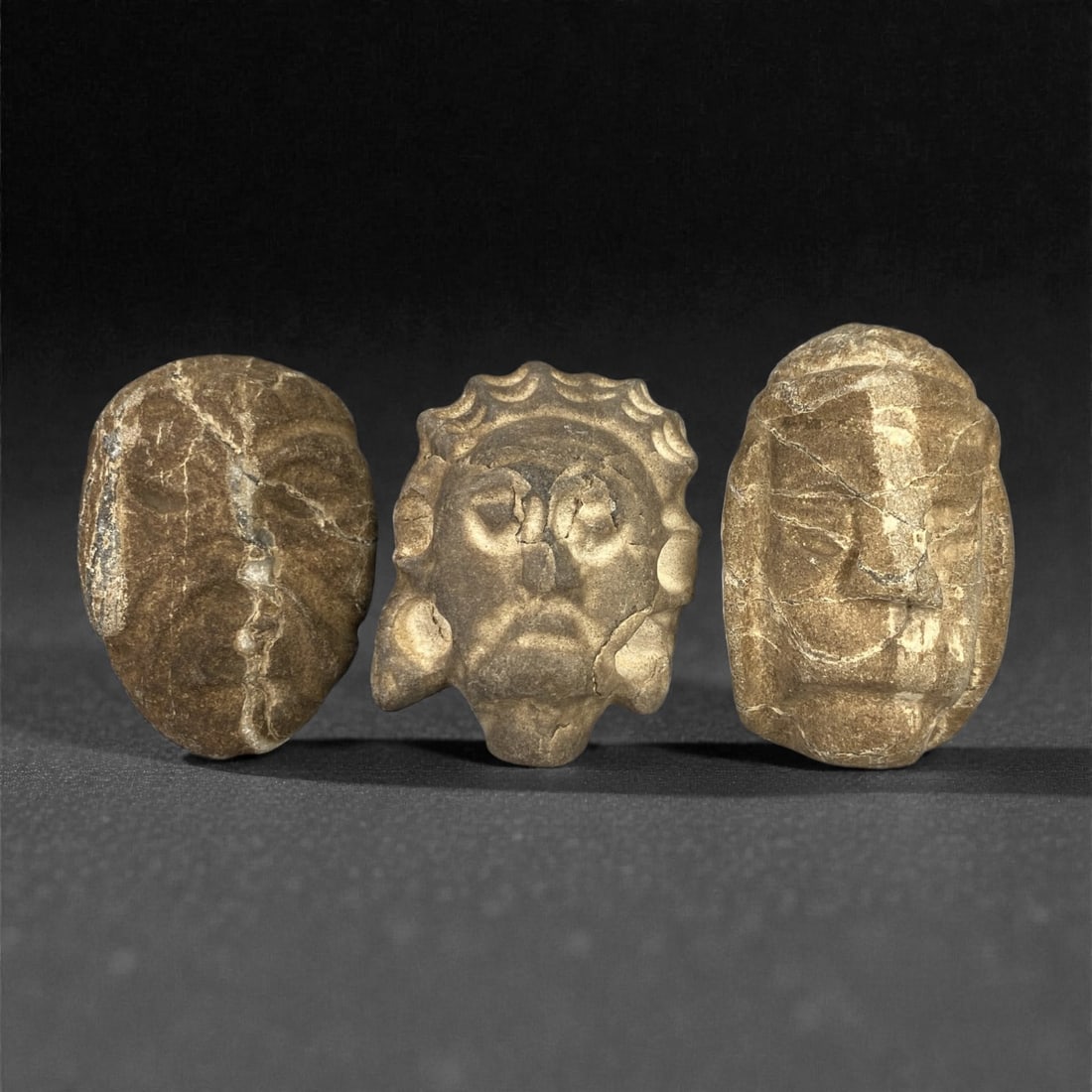 A Group of Three Ancient Stone Maskettes, 1st Millennium BCE, 21FN: Carved from stone with natural patination, mineral inclusions, and visible fissures, the three maskettes display distinct facial features. The leftmost example is ovoid, with a prominent vertical frac