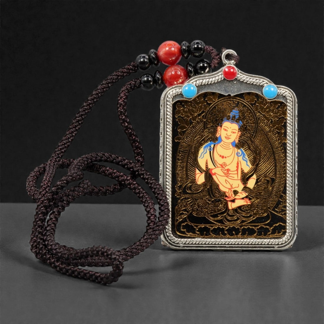 A Tibetan Metal and Painted Amulet, 20th Century, 48YC: The amulet is constructed with a rectangular metal frame housing a painted devotional image, suspended from a braided cord with applied beads. The central panel depicts a seated Buddhist figure shown