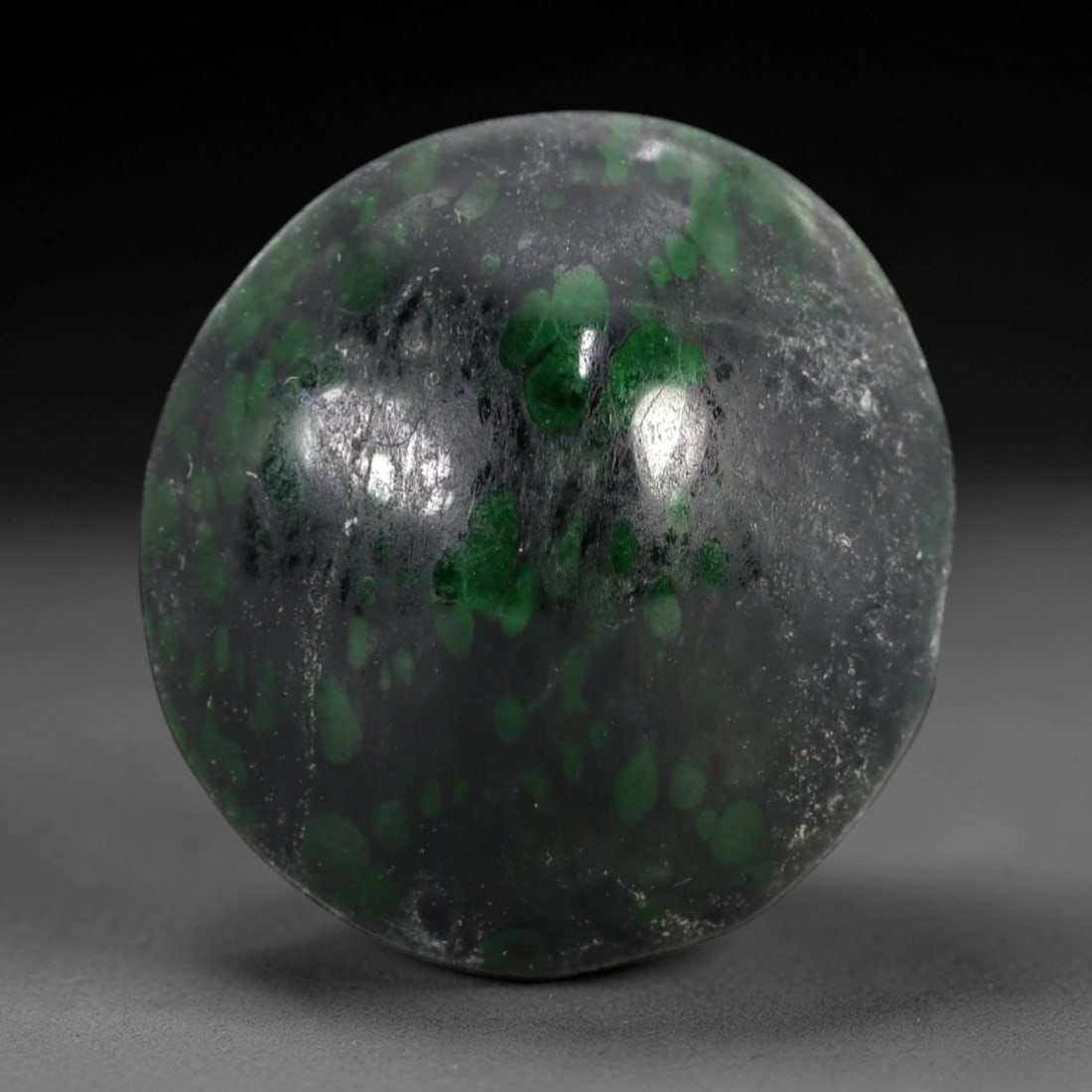 13.55 Carats Dark Matrix and Green Spot Maw Sit Sit Jade Cabochon - Natural Burmese Stone, 25AB: This maw sit sit jade cabochon is fashioned into a rounded, domed form with a highly polished surface. The material exhibits a dark, semi-translucent to opaque matrix, densely interspersed with distin