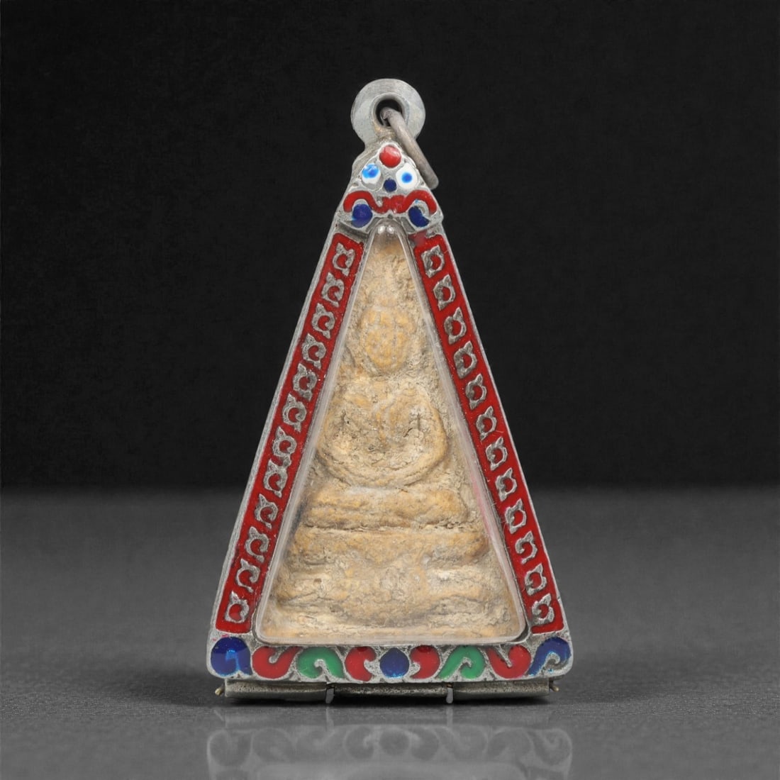 A Thai Clay Buddha Amulet in Triangular Silver Enamel Case, 20th Century, 93FN: Molded from light tan clay with visible surface patination and wear, the amulet depicts a seated Buddha in meditative posture. The reverse is plain, with uneven surface texture and age-related discolo