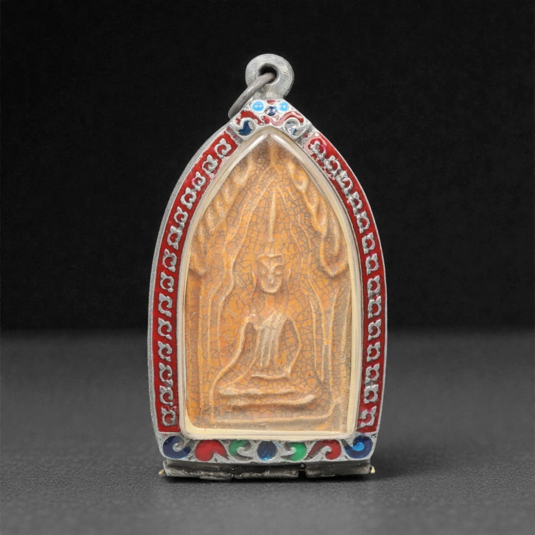 A Thai Clay Buddha Amulet in Silver Enamel Case, 20th Century, 88FN: Molded from light tan clay with visible craquelure and surface patination, the amulet depicts a seated Buddha with an ushnisha, set within a flame-shaped nimbus. The reverse is plain, with uneven surf