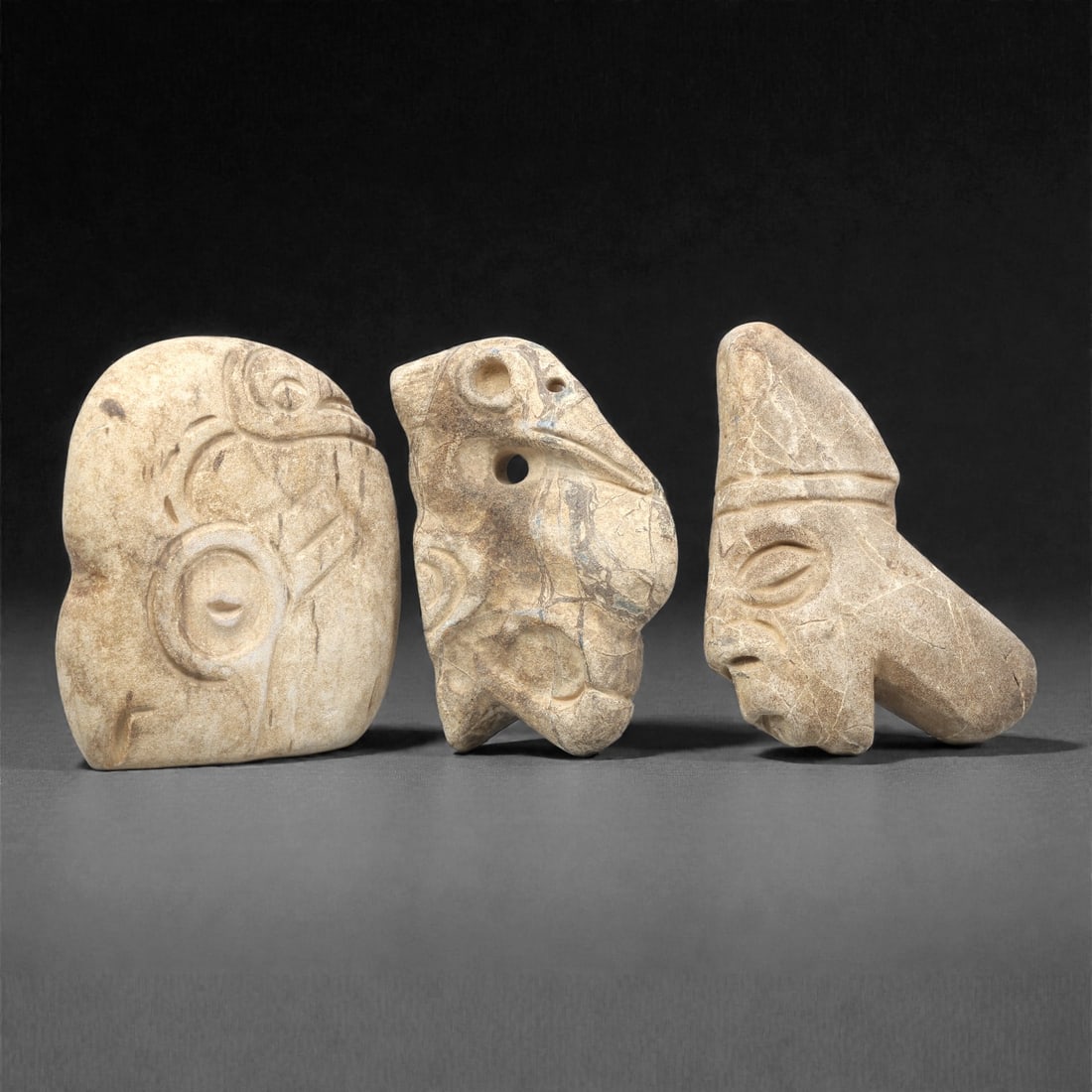 Three Pre-Columbian Stone Carvings, 10th-15th Century, 94ZG: The first carving is formed from light beige stone with natural veining, depicting a stylized head with a prominent circular earspool and incised facial motifs. The second is a zoomorphic pendant, car