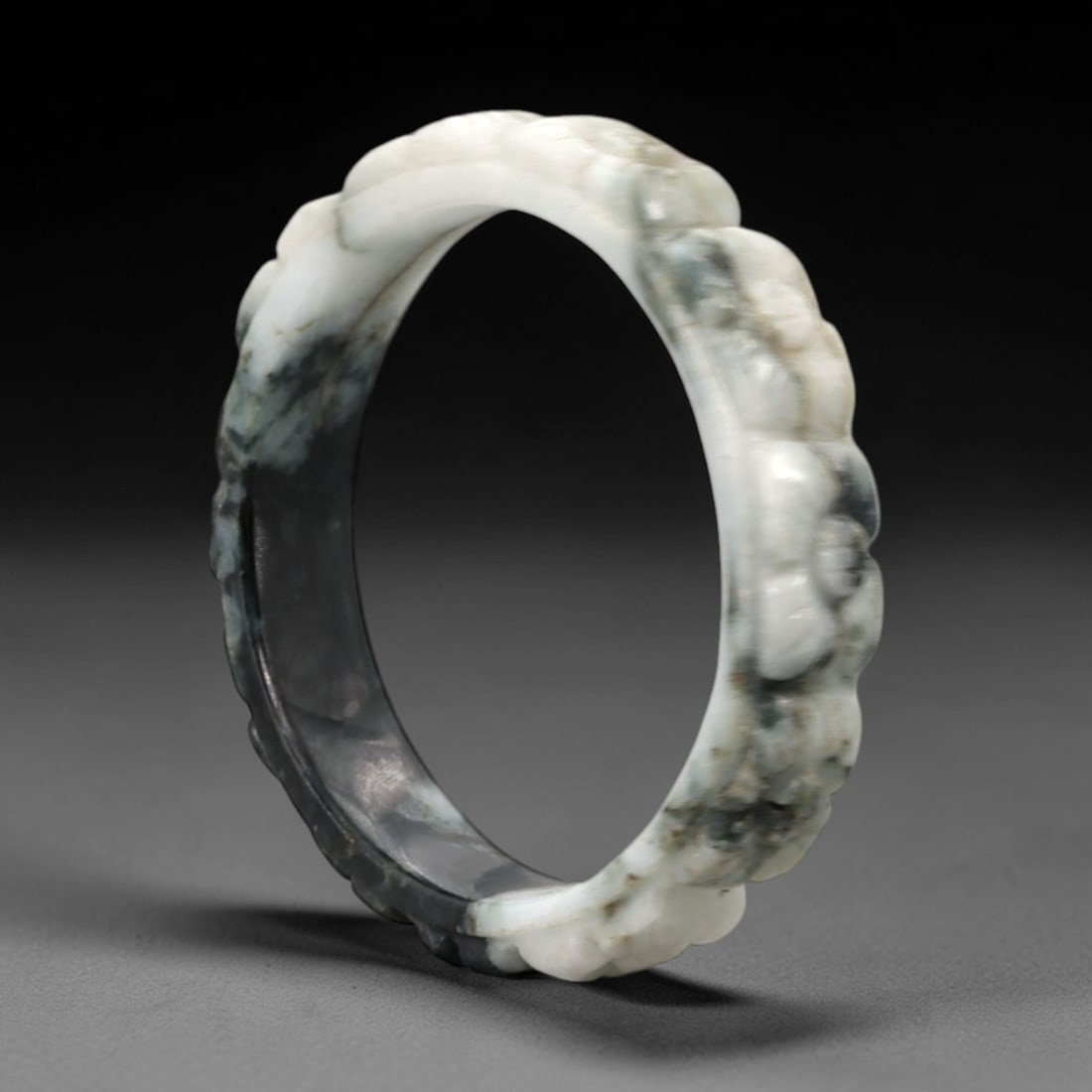 A Chinese Carved Jadeite Jade Bangle Bracelet, 20th Century, 63ZL: A fine jadeite bangle of flattened circular form, masterfully carved in high relief with a continuous band of stylized lingzhi fungus motifs. The stone exhibits a striking variegated palette, with a t