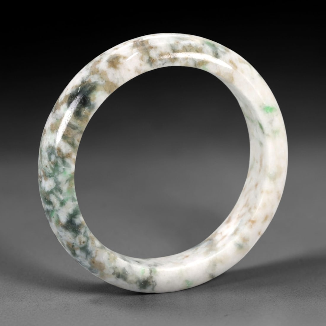 A Chinese Jadeite Jade Bangle Bracelet, 20th Century, 80ZK: Carved from jadeite jade with a pale white base and irregular mottling of green, brown, and dark grey inclusions, the bangle is formed as a continuous circular band with a rounded cross-section. The s