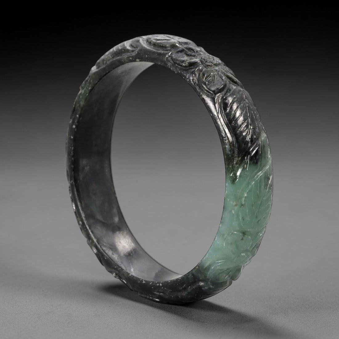 A Chinese Carved Jadeite Jade Bangle Bracelet, 20th Century, 64ZL: A substantial jadeite bangle of flattened circular form, masterfully carved in relief with a continuous band of floral blossoms and scrolling foliate motifs. The stone exhibits a striking variegated p