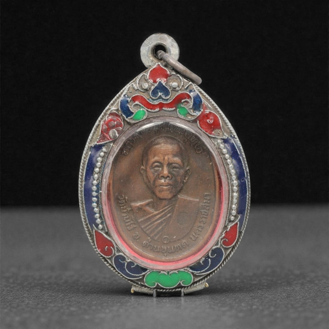 A Thai Monk Portrait Amulet Pendant, 20th Century, 23FP: This teardrop-shaped pendant features a copper alloy core, encased in an ornate silver frame with a transparent protective cover, measuring 5.1 x 3.3 cm (2 x 1 5/16 in.). The obverse displays a detail