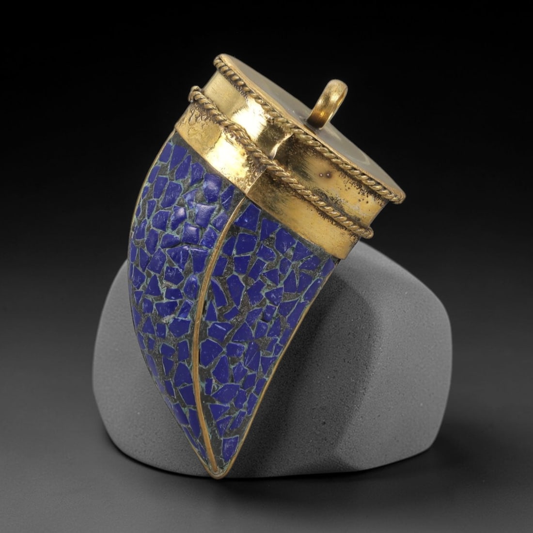 A Tibetan Gilt-Metal and Lapis Lazuli Mosaic Gau Pendant, 20th Century, 87ZL: This Tibetan gau (prayer amulet) is of distinctive horn-shaped form, crafted from a gilt-brass alloy with a rich, aged patina. The curved body is adorned with a vibrant mosaic of deep blue lapis lazul