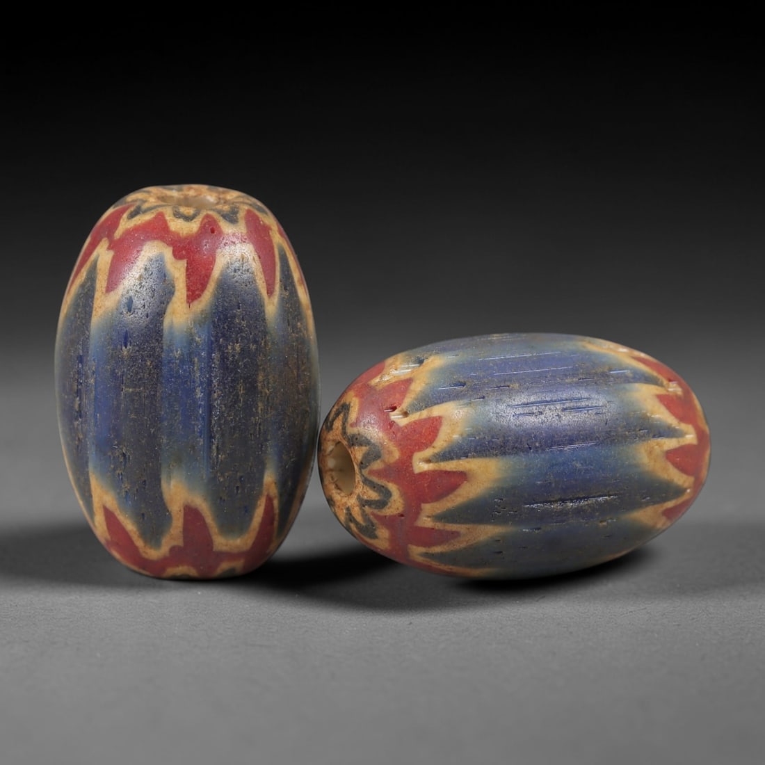 A Pair of Venetian Glass Chevron Beads, 18th-19th Century, 76AC: Crafted from translucent glass with a pale yellow core, each bead is decorated with a chevron pattern of alternating deep blue and vibrant red layers, radiating from the central perforation to the out