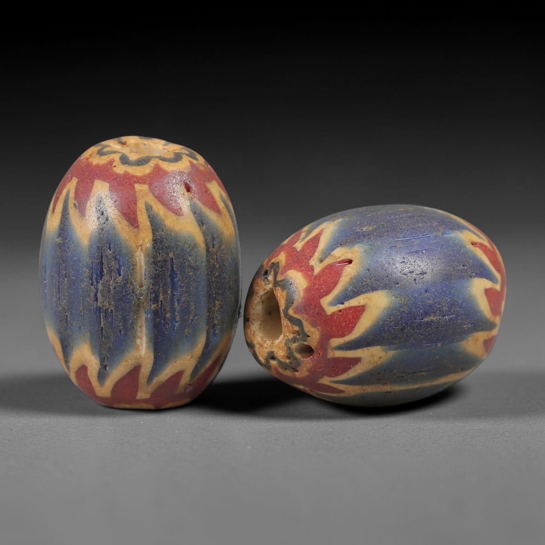 A Pair of Venetian Glass Chevron Beads, 18th-19th Century, 77AC: Crafted from translucent glass with a pale yellow core, each bead is decorated with a chevron pattern of alternating deep blue and vibrant red layers, radiating from the central perforation to the out