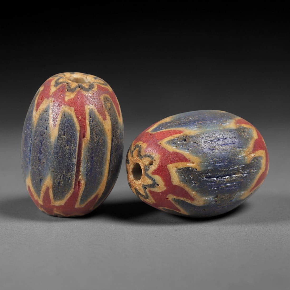 A Pair of Venetian Glass Chevron Beads, 18th-19th Century, 85AC: Crafted from translucent glass with a pale yellow core, each bead is decorated with a chevron pattern of alternating deep blue and vibrant red layers, radiating from the central perforation to the out