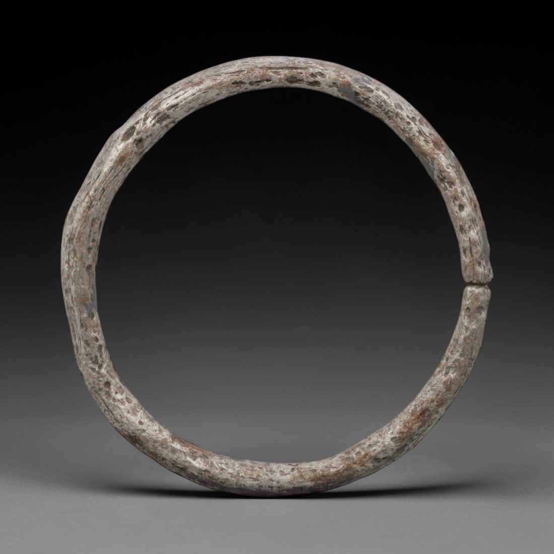 A Celtic Iron Bracelet, 2nd Century BCE - 1st Century CE, 2FV: Forged from iron with a thick, rounded section, the bracelet is formed as a near-complete circle with a narrow terminal gap. The surface exhibits a pitted, silvery-grey patina with extensive corrosion