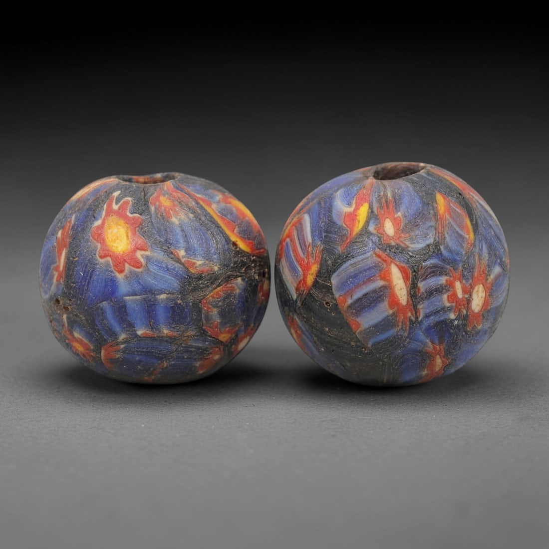 A Pair of Venetian Millefiori Glass Beads, 19th-20th Century, 70AD: Fabricated from opaque dark blue glass, each bead is formed as a sphere with a central drilled perforation. The surface is decorated with characteristic millefiori canes, featuring floral and leaf-lik
