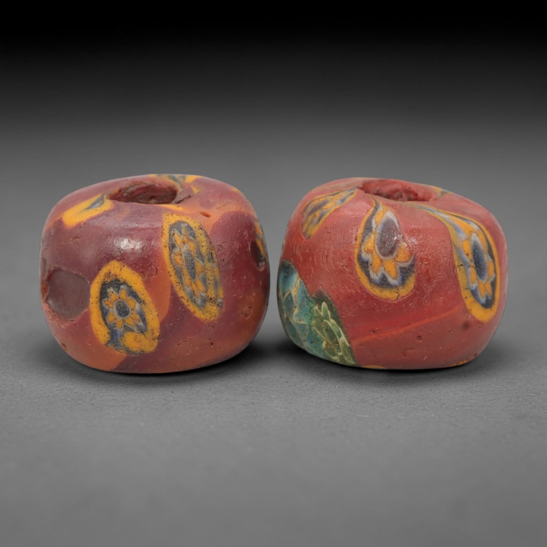 A Pair of Venetian Millefiori Glass Beads, 19th-20th Century, 72AD: Fabricated from opaque red glass, each bead is formed as a short cylinder with a central drilled perforation. The surface is decorated with characteristic millefiori canes, featuring eye-like motifs w