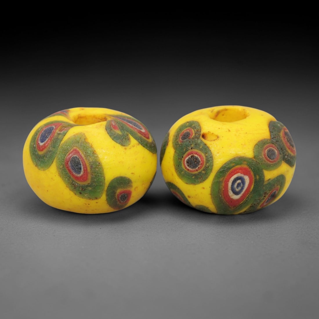 A Pair of Venetian Millefiori Glass Beads, 19th-20th Century, 60AD: Fabricated from opaque yellow glass, each bead is formed as a short cylinder with a central drilled perforation. The surface is decorated with characteristic millefiori canes, featuring concentric cir