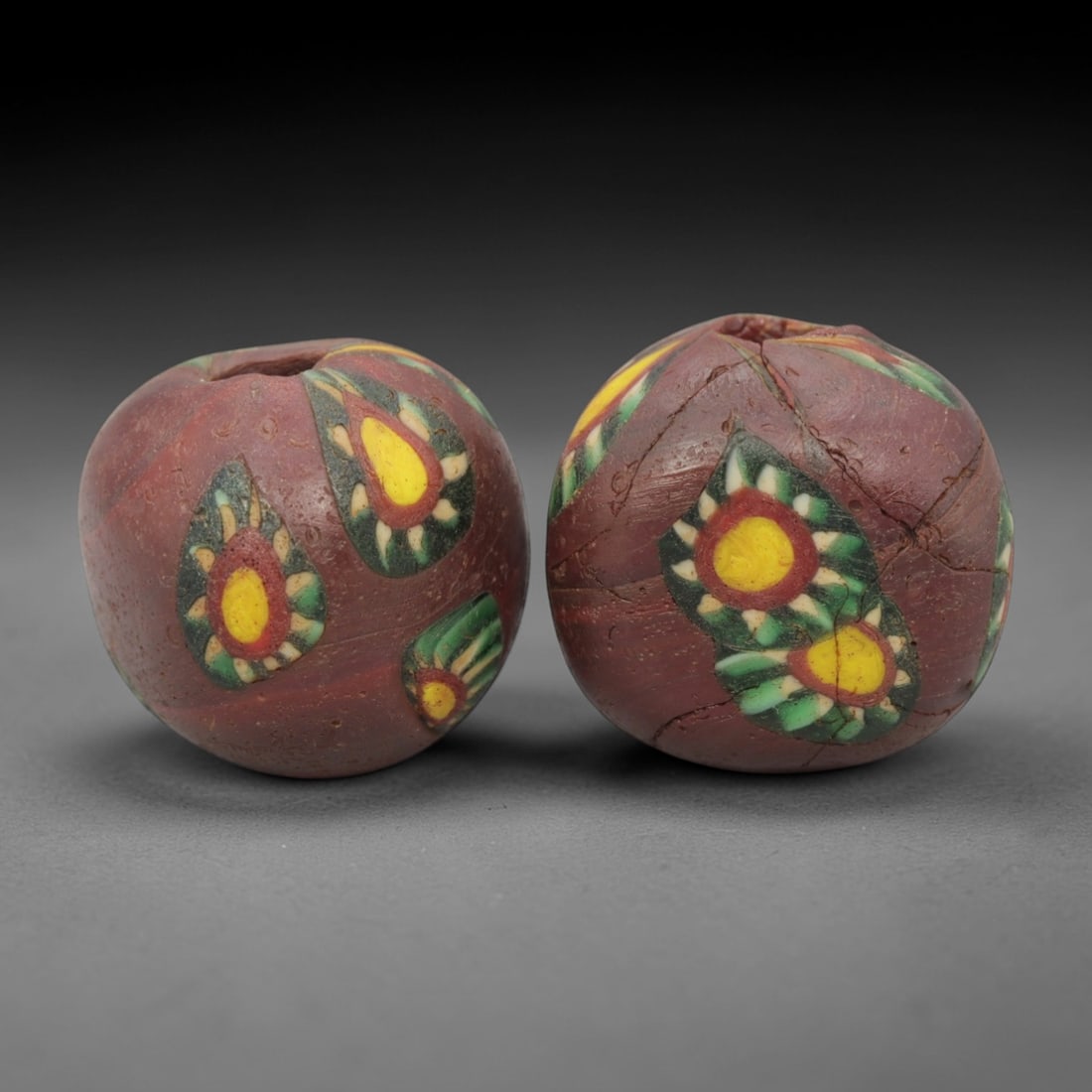 A Pair of Venetian Millefiori Glass Beads, 19th-20th Century, 61AD: Crafted from opaque brown glass, each bead is formed as a sphere with a central drilled perforation. The surface is decorated with characteristic millefiori canes, featuring circular floral or eye-lik