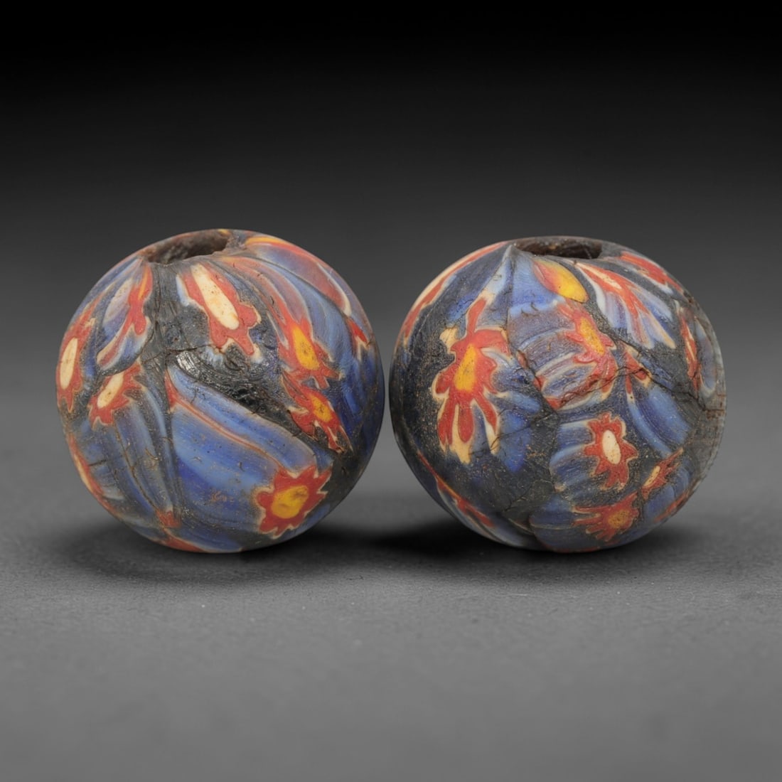 A Pair of Venetian Millefiori Glass Beads, 19th-20th Century, 57AD: Fabricated from opaque dark blue glass, each bead is formed as a sphere with a central drilled perforation. The surface is decorated with characteristic millefiori canes, featuring floral and leaf-lik