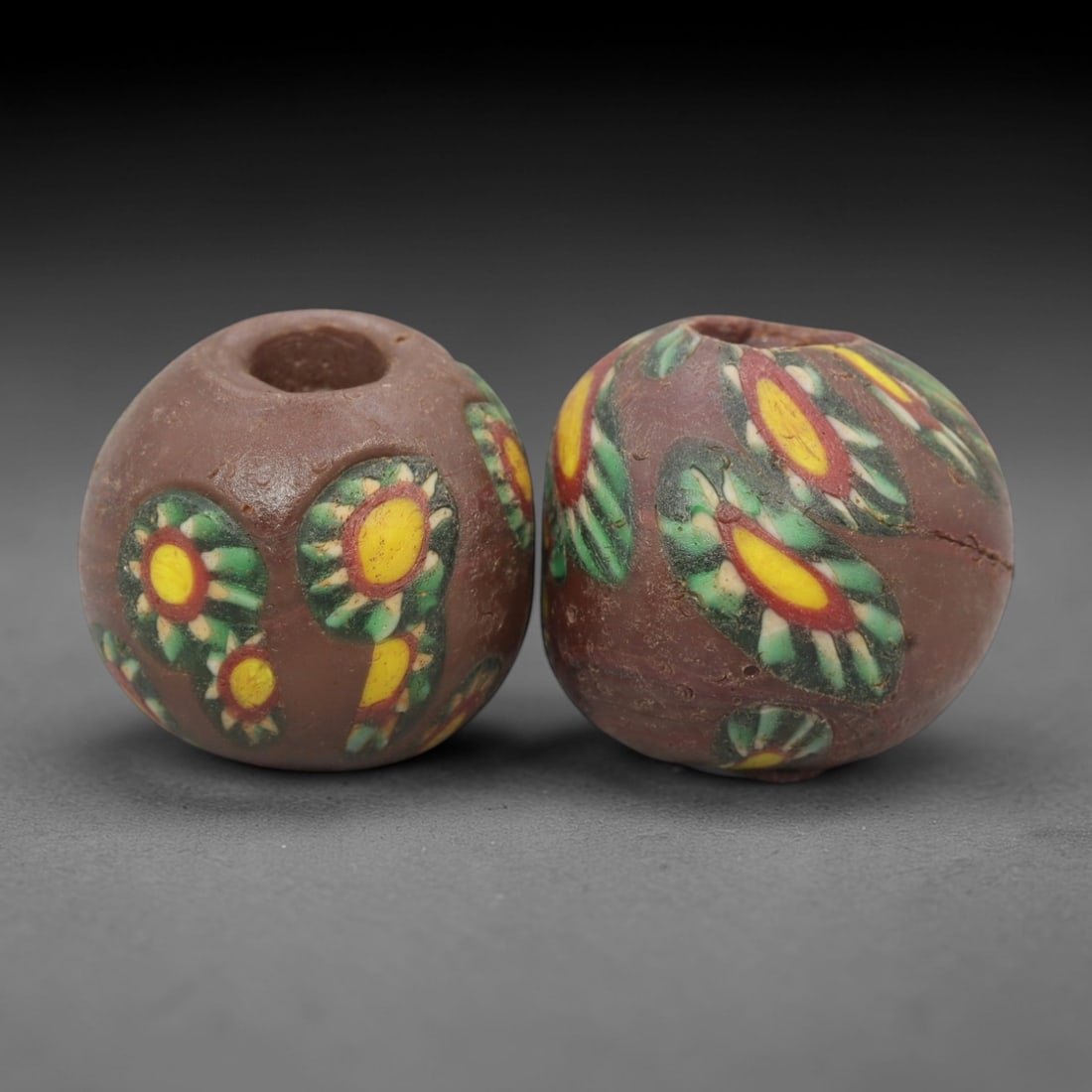 A Pair of Venetian Millefiori Glass Beads, 19th-20th Century, 73AD: Fabricated from opaque brown glass, each bead is formed as a sphere with a central drilled perforation. The surface is decorated with characteristic millefiori canes, featuring circular floral or eye-