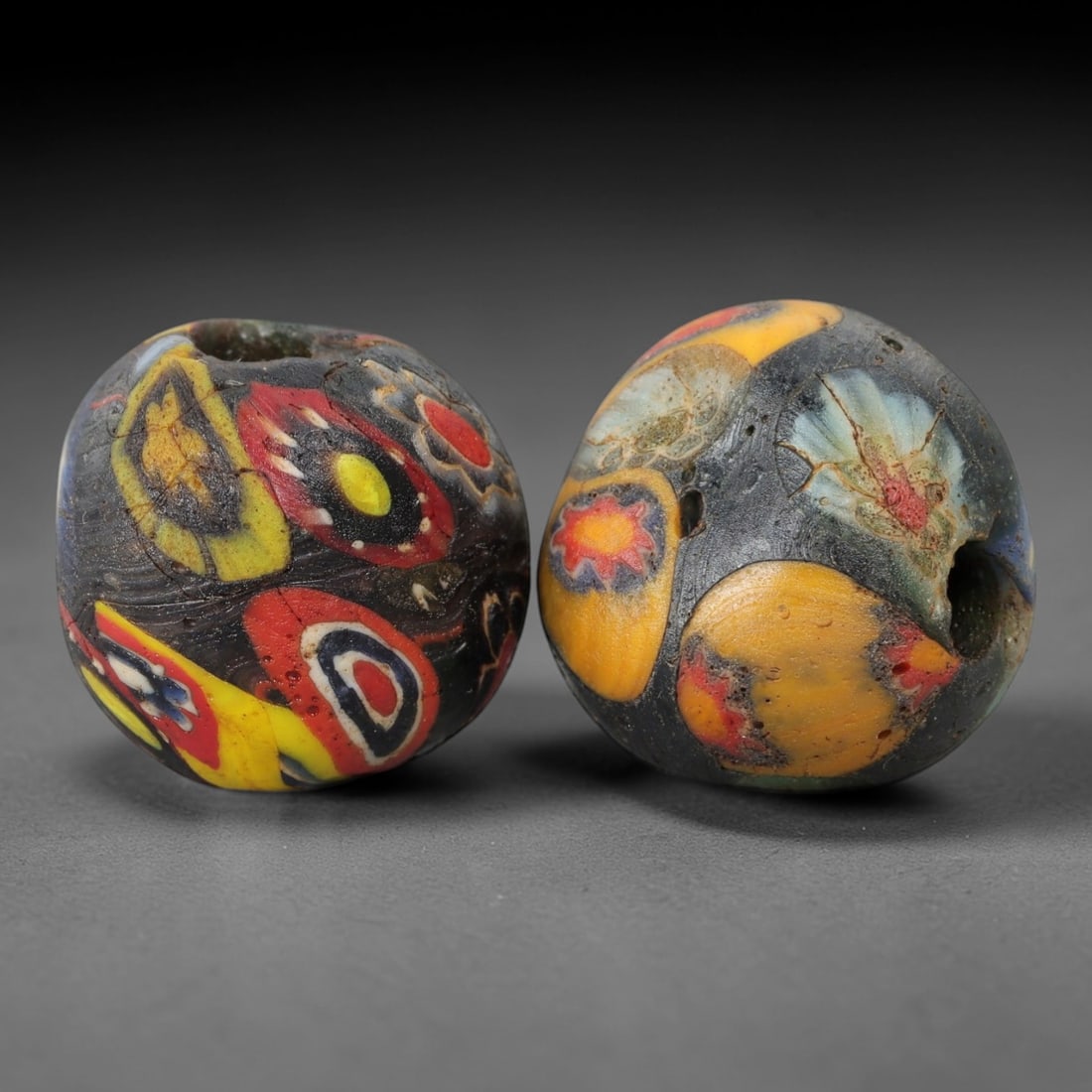 A Pair of Venetian Glass Trade Beads, 19th-20th Century, 83ZM: Formed as spherical beads, each is crafted from dark translucent glass with a central perforation for suspension. The surface is decorated with multiple polychrome eye and floral motifs, featuring red