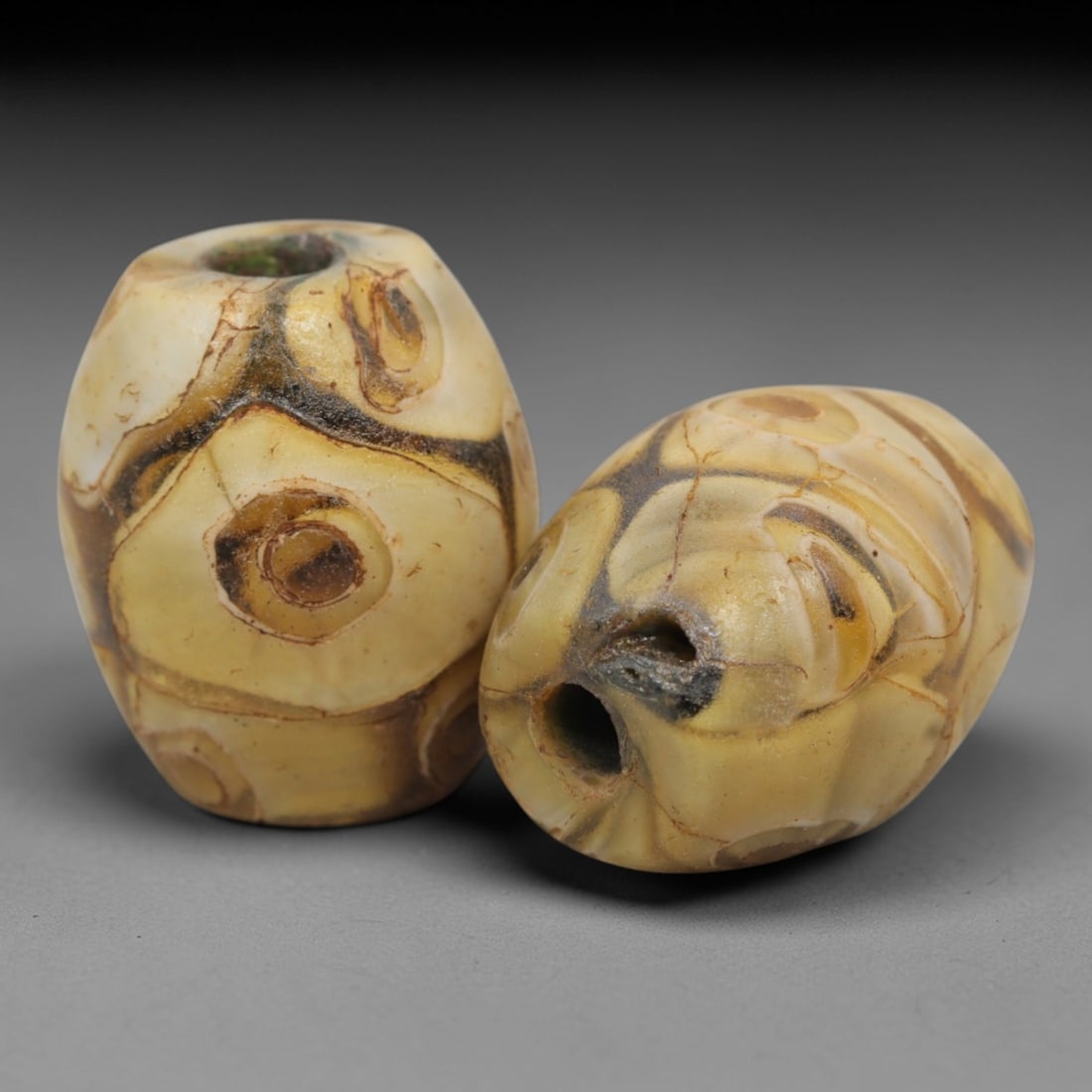 A Pair of Venetian Glass Trade Beads, 19th-20th Century, 23ZM: Formed as barrel-shaped beads, each is crafted from translucent yellowish-brown glass with a central perforation for suspension. The surface is decorated with multiple concentric circular eye motifs,