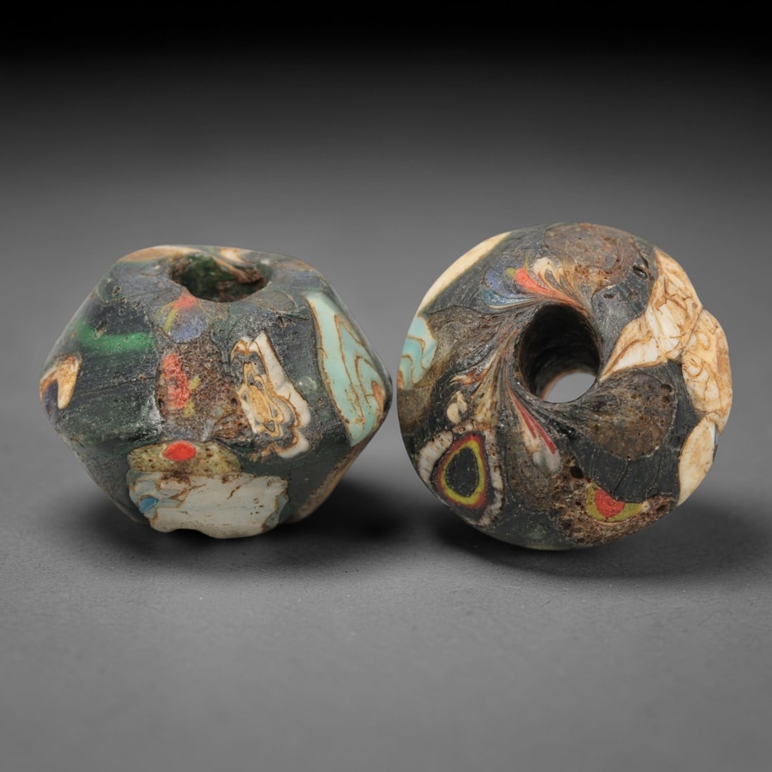 A Pair of Venetian Glass Trade Beads, 19th-20th Century, 86ZM: Formed as faceted and spherical beads, each is crafted from dark translucent glass with a central perforation for suspension. The surface displays embedded, fragmented inclusions of opaque colored gla