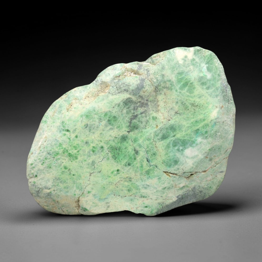 A Burmese Jadeite Jade Rough, 20th Century, 59FS: A piece of Burmese jadeite jade rough, with a pale greenish-white matrix and vibrant green mineral inclusions. The rough is shaped as an irregular, flattened form, with visible natural fractures and a